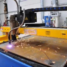 Proces cięcia plazmowego blachy stalowej na maszynie CNC, z widocznymi iskrami i śladami cięcia. Żółto-niebieska maszyna z napisem 'ajan' w hali produkcyjnej.