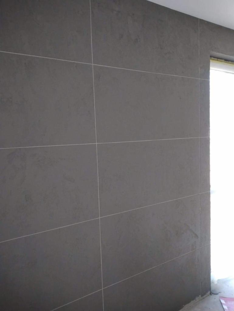 Ściana wykończona szarymi płytami z delikatną teksturą i jasnymi fugami, widok z dołu, fragment okna w tle.