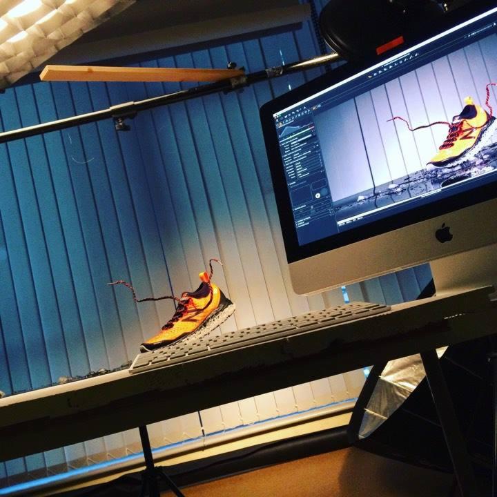 Pomarańczowo-żółty but sportowy zawieszony w powietrzu podczas sesji zdjęciowej, widoczny monitor iMac z oprogramowaniem do edycji graficznej, oświetlenie studyjne w tle.