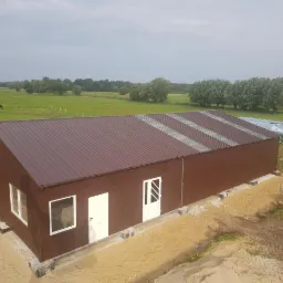 Gotowa hala magazynowa z brązową elewacją i trapezowym dachem z świetlikami, widok z zewnątrz na tle zielonego pola i lasu, częściowo zagospodarowany teren wokół budynku.