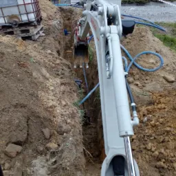 Wykop pod rury, wykonany minikoparką, widoczne ułożone rury w wykopie oraz zbiornik IBC na palecie w tle.