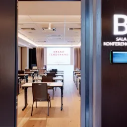 Sala konferencyjna z rzędami stołów i krzeseł, widok zza otwartych drzwi, na ścianie tabliczka z napisem 'Sala Konferencyjna' i numerem 'B' oraz panel dotykowy.