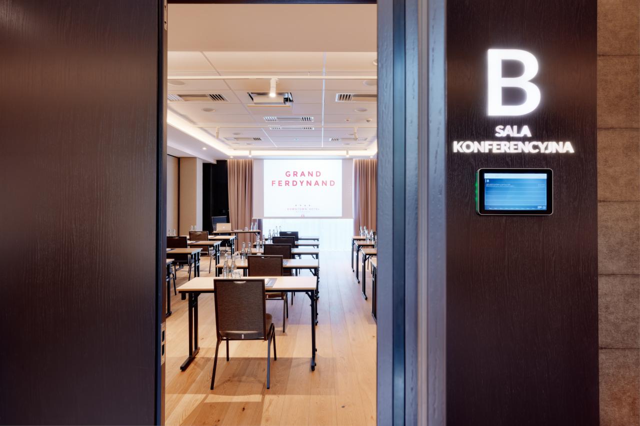 Sala konferencyjna z rzędami stołów i krzeseł, widok zza otwartych drzwi, na ścianie tabliczka z napisem 'Sala Konferencyjna' i numerem 'B' oraz panel dotykowy.
