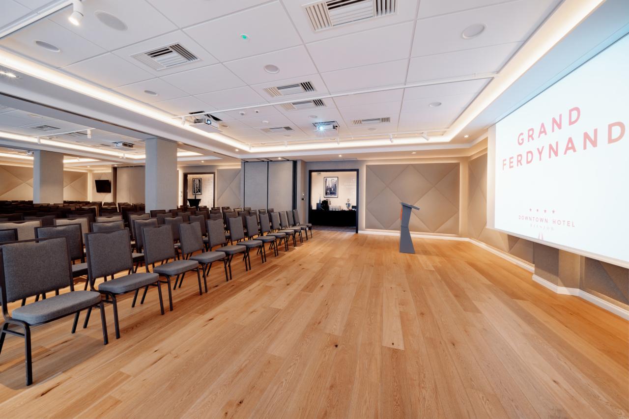 Sala konferencyjna z rzędami krzeseł, drewnianą podłogą i ekranem z napisem Grand Ferdynand Downtown Hotel.