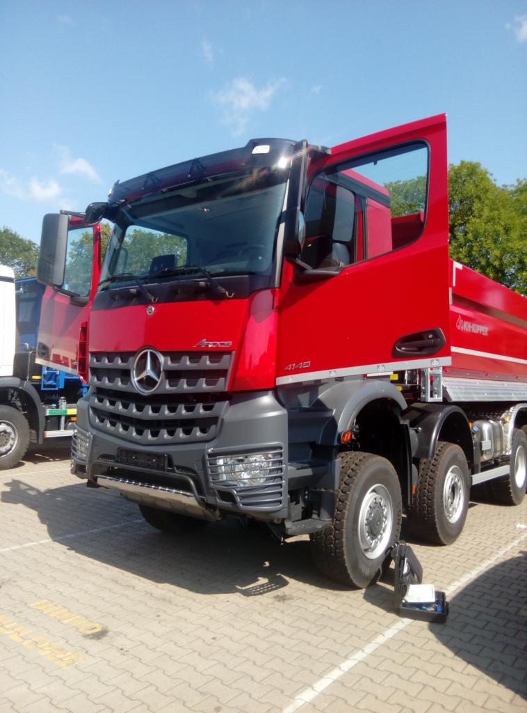 Czerwona ciężarówka Mercedes-Benz Arocs 4145 z otwartymi drzwiami kabiny, widoczna na placu z kostki brukowej, z innym pojazdem w tle.