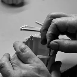 Czarno-białe zbliżenie rąk montera wkręcających śrubę w plastikową obudowę; w tle leżą śruby i wkręty.