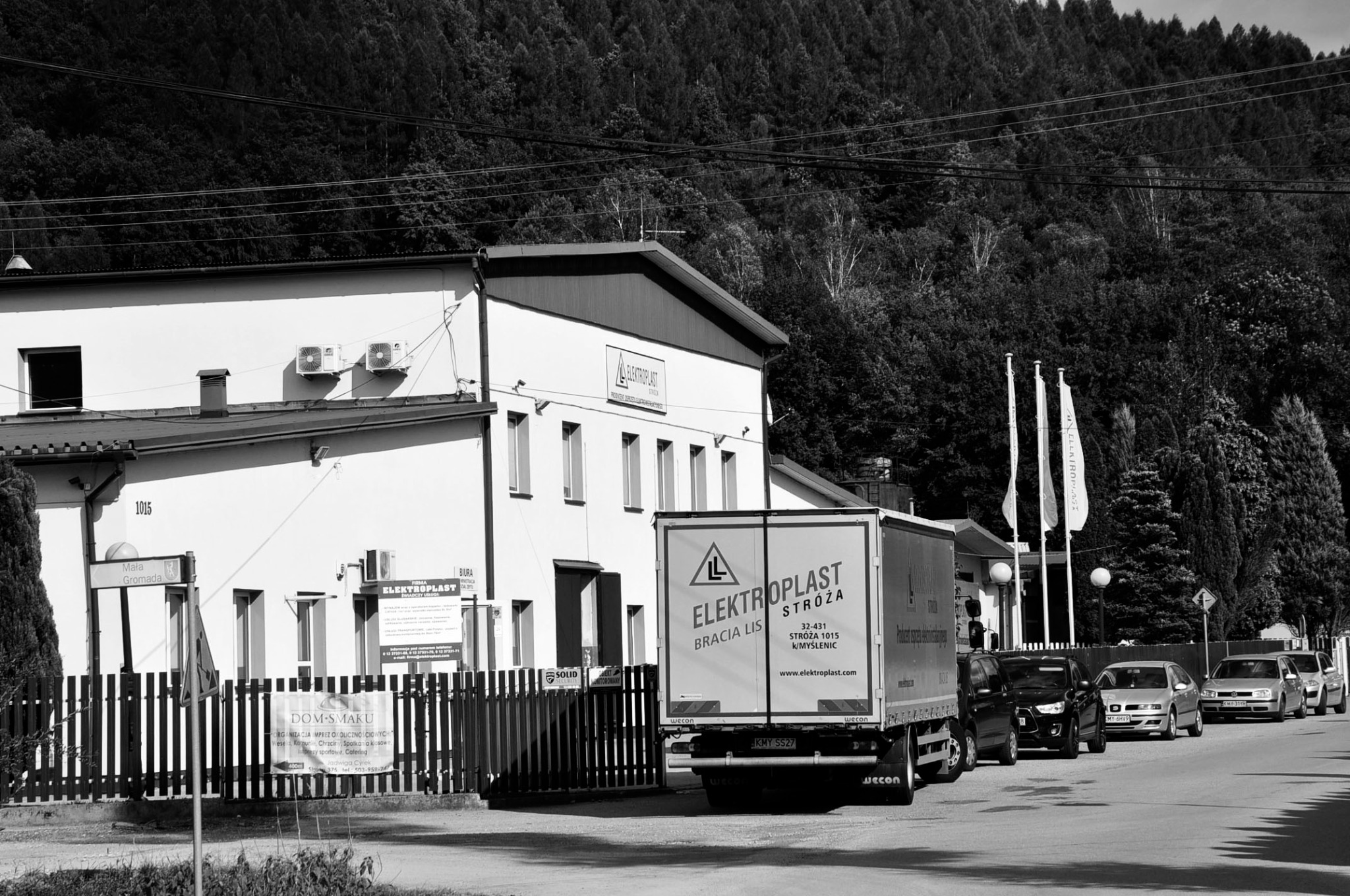 Czarno-białe zdjęcie budynku firmy Elektroplast Stroża z zaparkowanym samochodem dostawczym tej firmy na pierwszym planie, w tle zalesione wzgórze.
