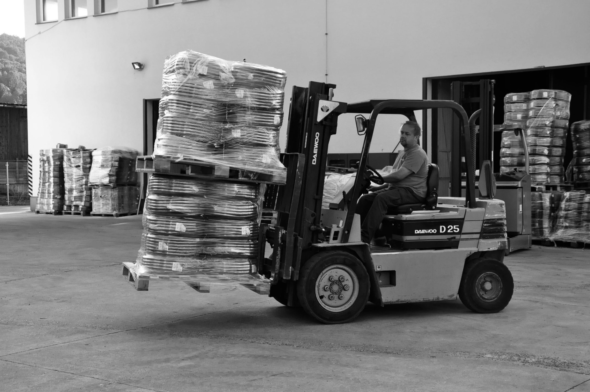 Czarno-białe zdjęcie wózka widłowego Daewoo D25 z operatorem, przewożącego palety z towarem owiniętym folią stretch na tle magazynu.