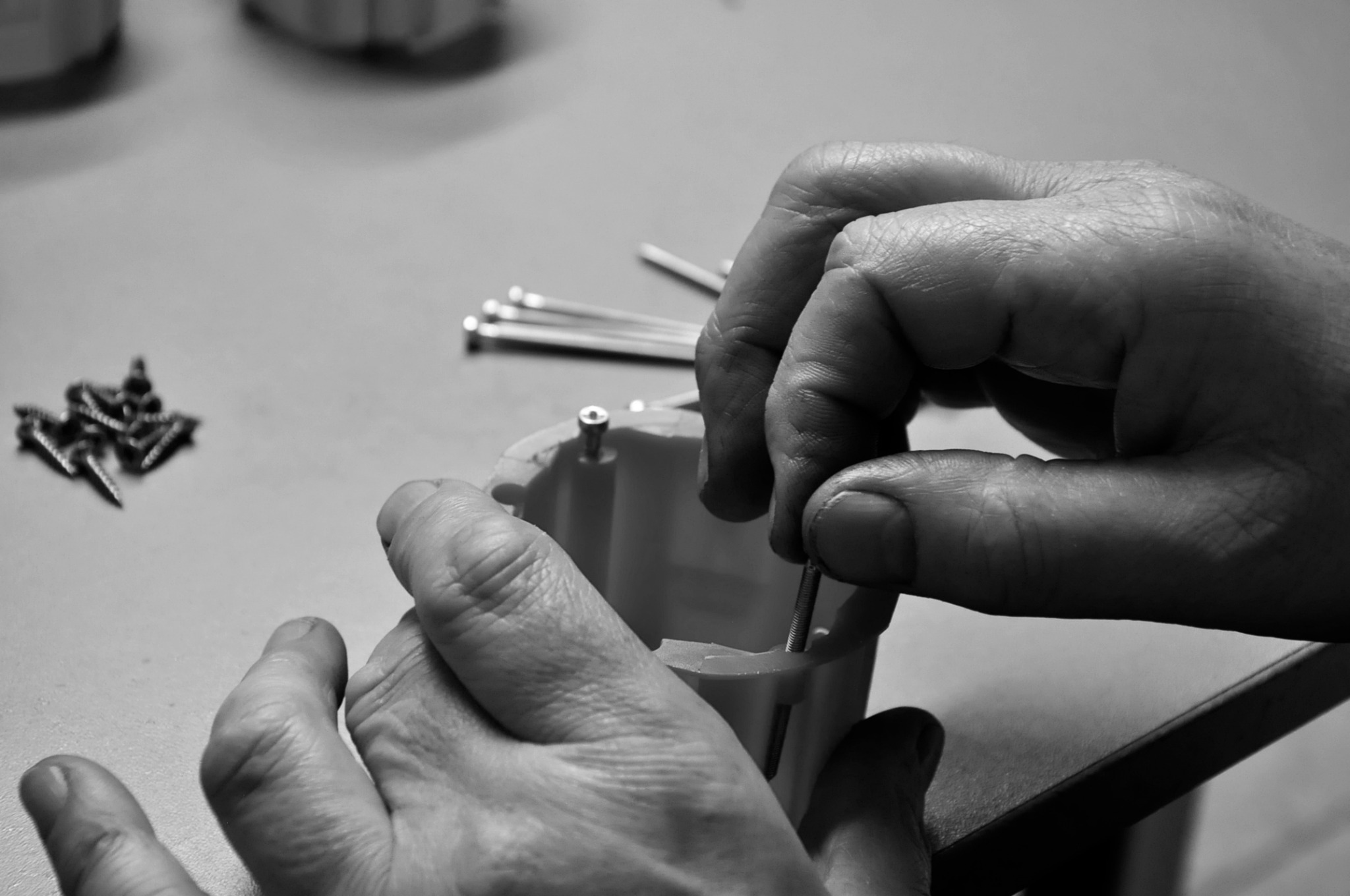 Czarno-białe zbliżenie rąk montera wkręcających śrubę w plastikową obudowę; w tle leżą śruby i wkręty.