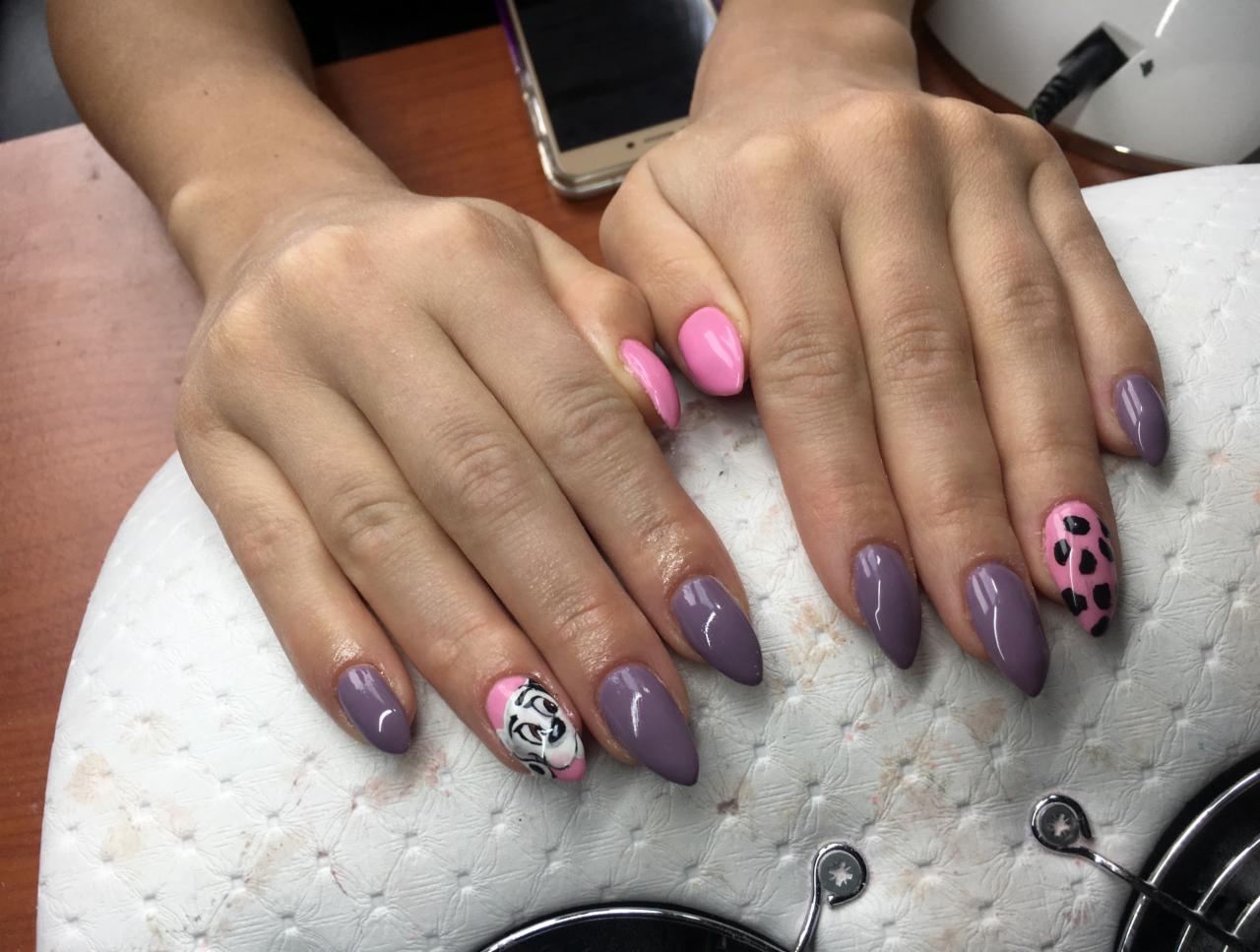 Dłonie z pomalowanymi paznokciami w odcieniach fioletu, różu i z wzorami: panterka i postać z bajki na tle białej podkładki.