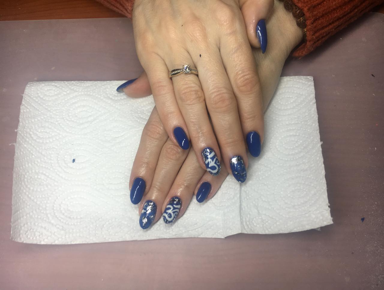 Dłonie kobiety z ciemnoniebieskimi paznokciami, zdobionymi srebrnymi drobinkami i białym wzorem na kilku paznokciach, pierścionek na palcu serdecznym, na białym podkładzie.