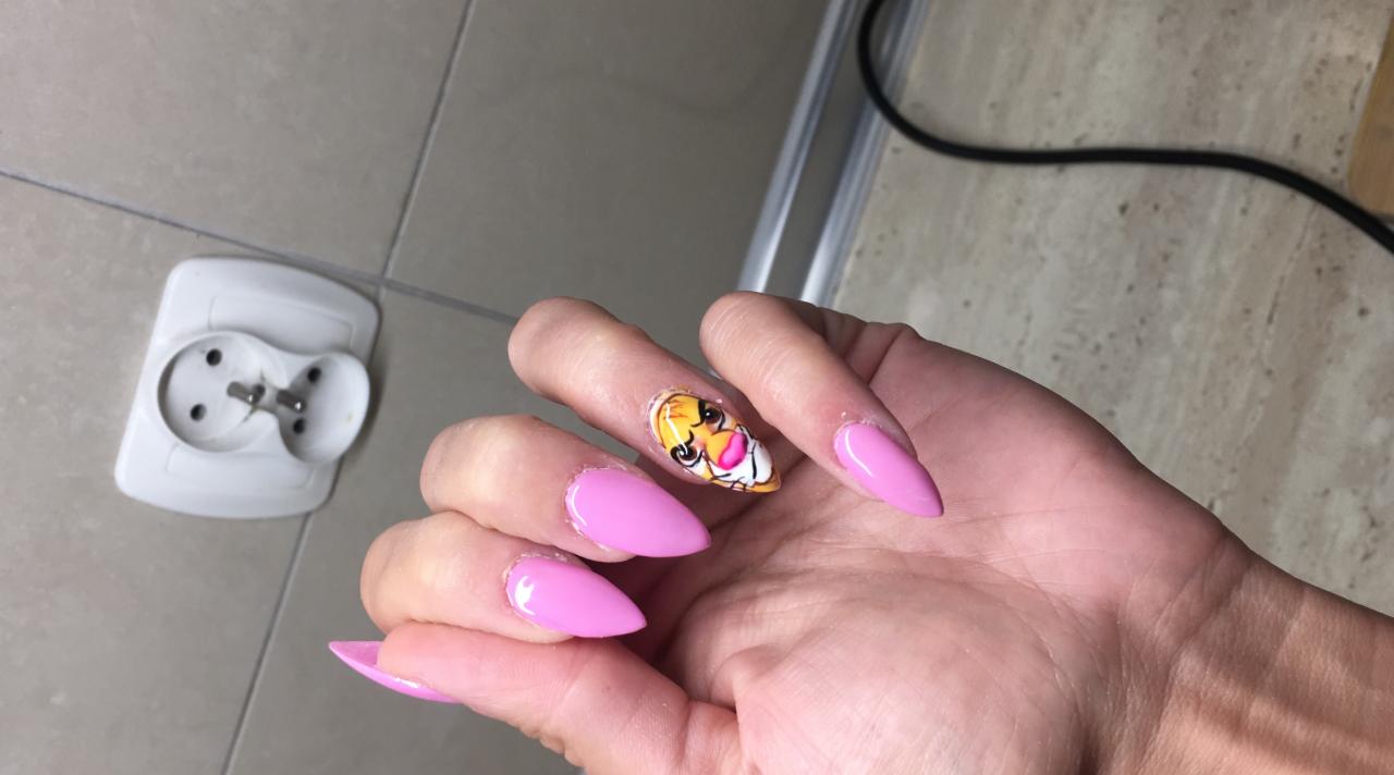 Dłoń z pomalowanymi na różowo paznokciami w kształcie migdała i paznokciem akcentującym z rysunkiem postaci z kreskówki, widoczna na tle łazienkowej ściany z gniazdkiem.