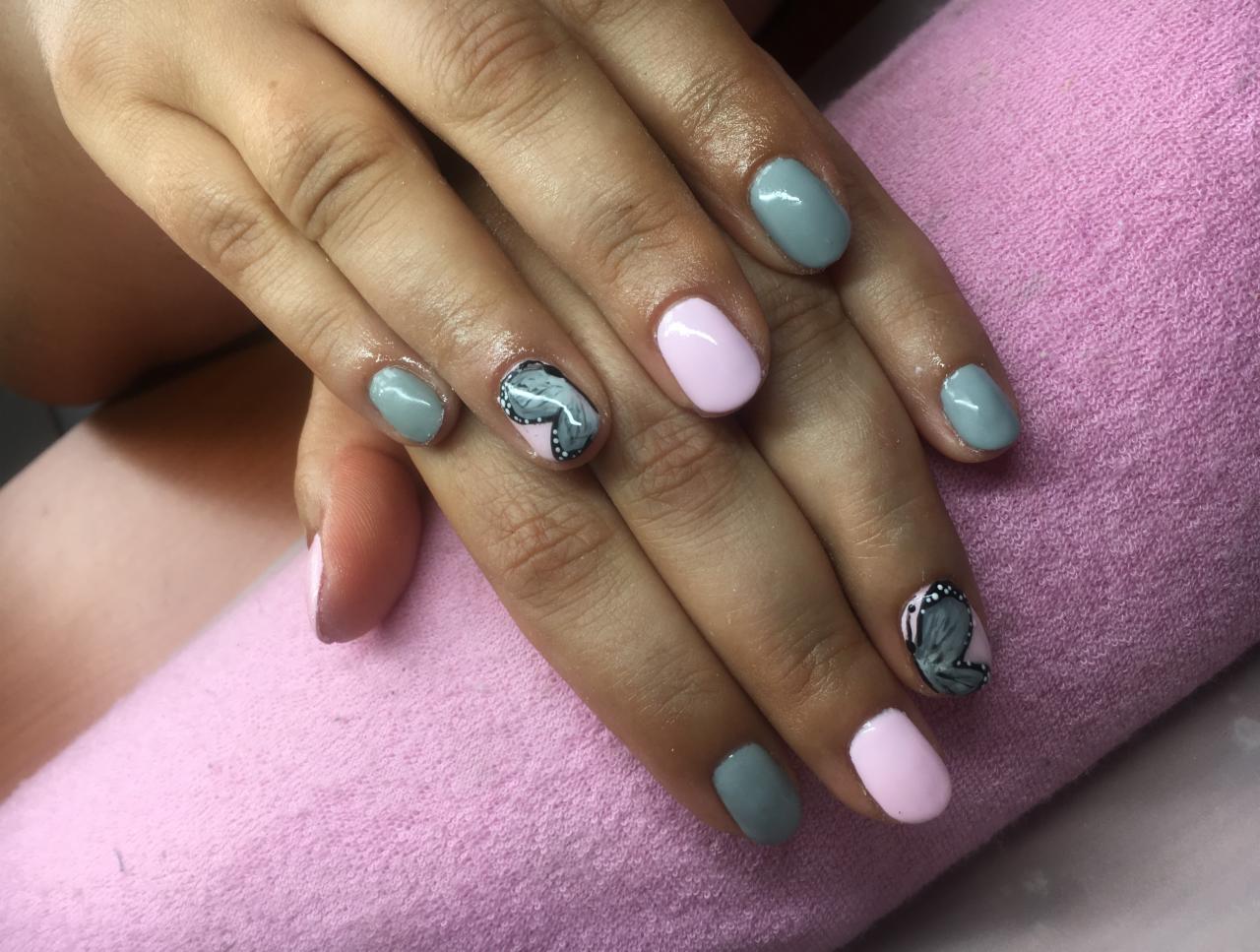 Dłonie z pomalowanymi paznokciami w odcieniach szarości i różu, na jednym z paznokci widoczny motyl na jasnoróżowym tle, ułożone na różowym ręczniku.