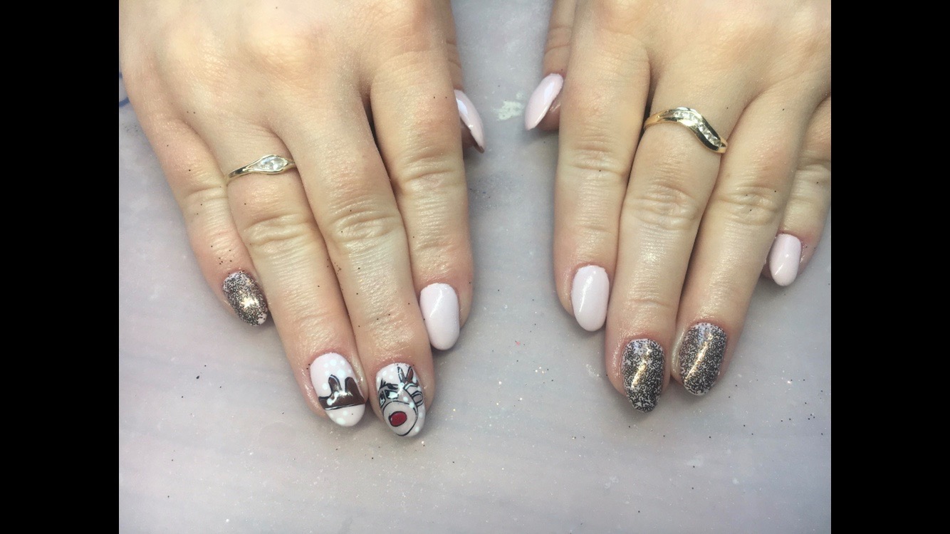 Dłonie z jasnoróżowym i brokatowym lakierem, z reniferem na paznokciu wskazującym, ozdobione pierścionkami na palcach serdecznych.