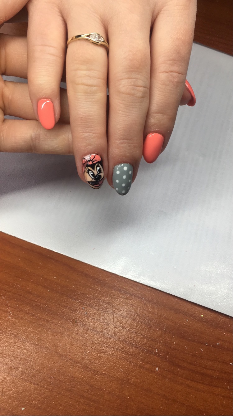 Dłoń z pomalowanymi paznokciami w odcieniach koralu i szarości, z wzorem Myszki Minnie i białymi kropkami, ozdobiona złotym pierścionkiem z oczkiem.