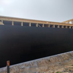 Nor House - Wznoszenie ściany zewnętrznej budynku z drewnianą konstrukcją szkieletową i czarną izolacją, widoczny fragment fundamentu i słupka geodezyjnego.