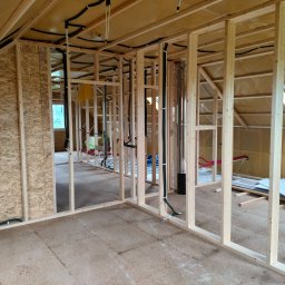 Nor House - Wnętrze w trakcie budowy: drewniany szkielet konstrukcji, widoczne instalacje elektryczne i rury grzewcze, surowa podłoga z płyty OSB.