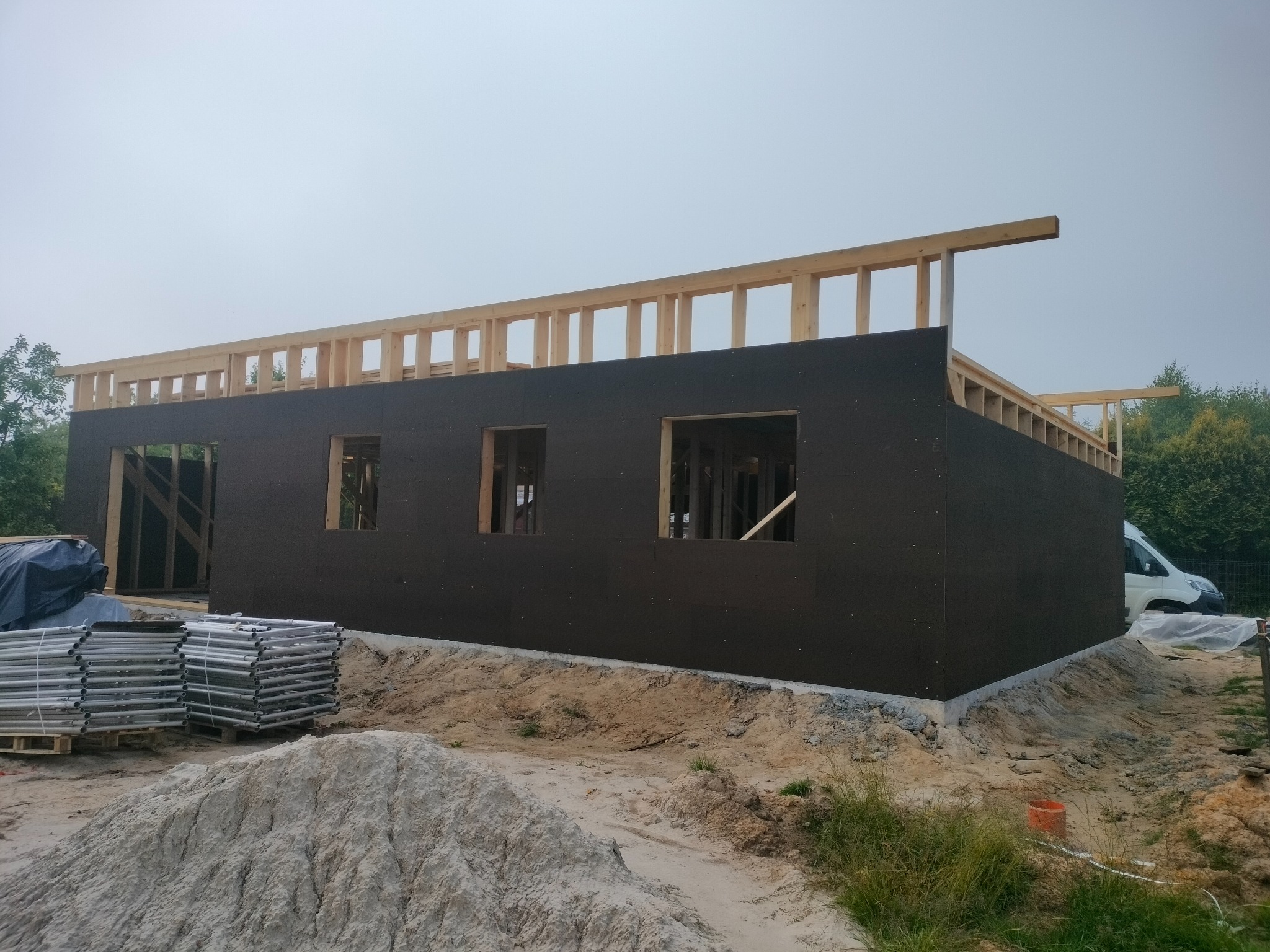 Budowa domu: surowa bryła z ciemną elewacją i drewnianą konstrukcją szkieletową dachu, widoczne rusztowania i hałda piasku.
