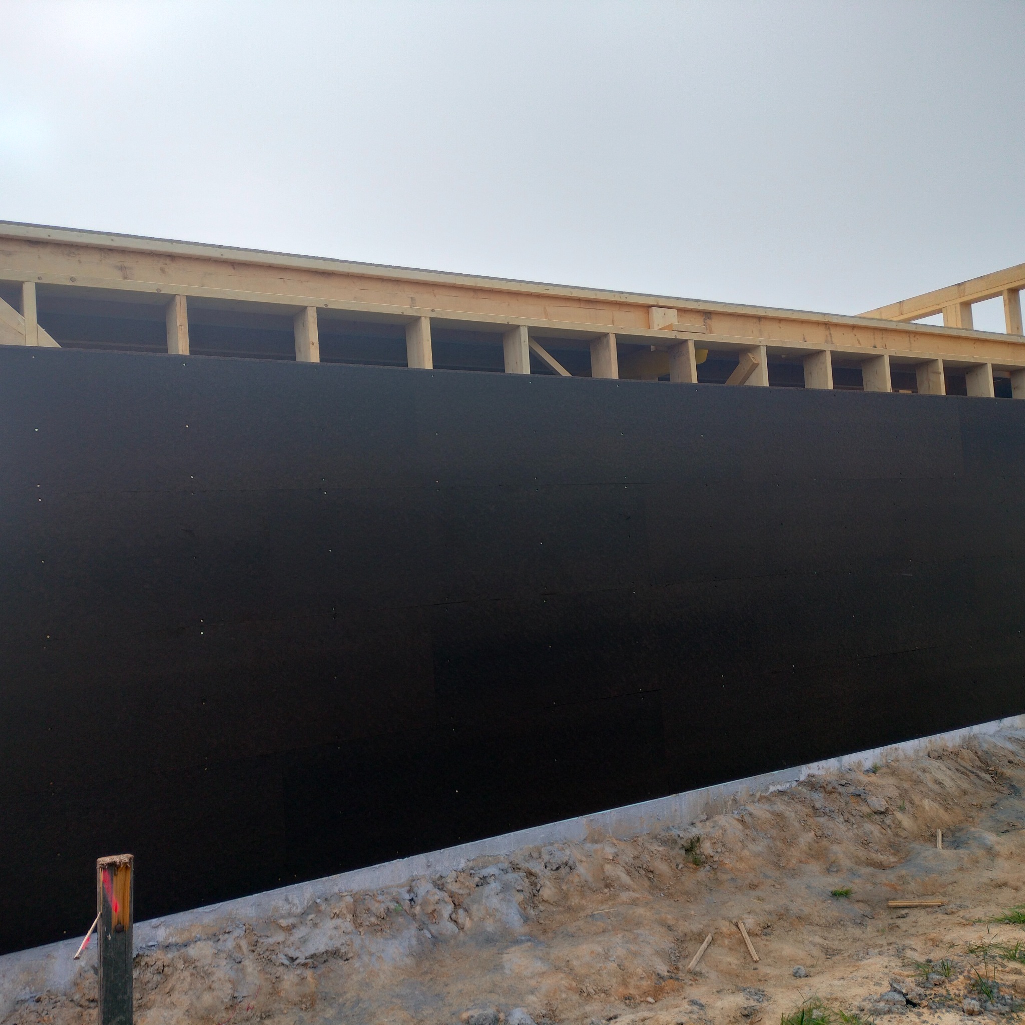 Wznoszenie ściany zewnętrznej budynku z drewnianą konstrukcją szkieletową i czarną izolacją, widoczny fragment fundamentu i słupka geodezyjnego.