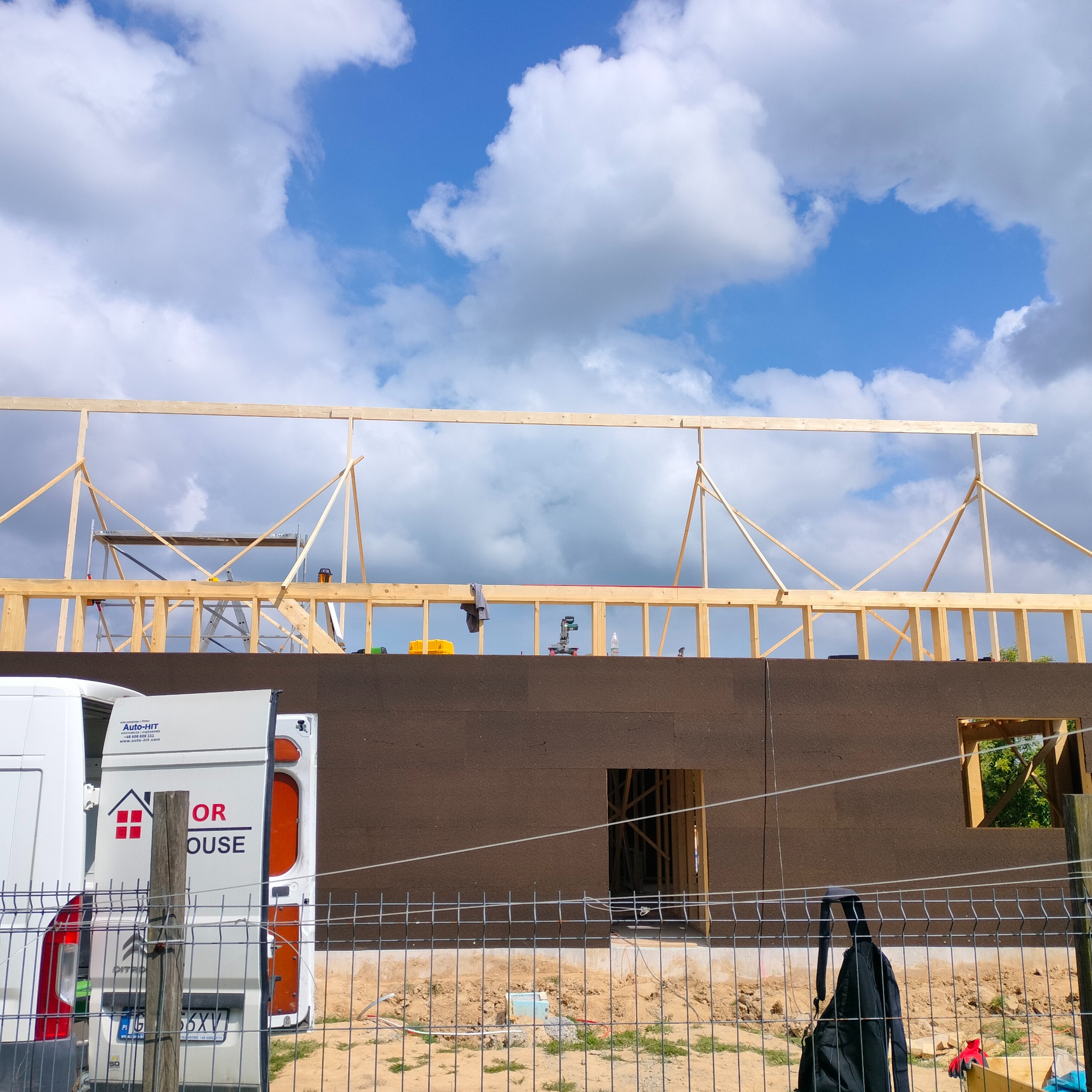 Konstrukcja drewniana szkieletu domu z widoczną izolacją ścian, na tle błękitnego nieba z chmurami, z zaparkowanym samochodem dostawczym firmy budowlanej.