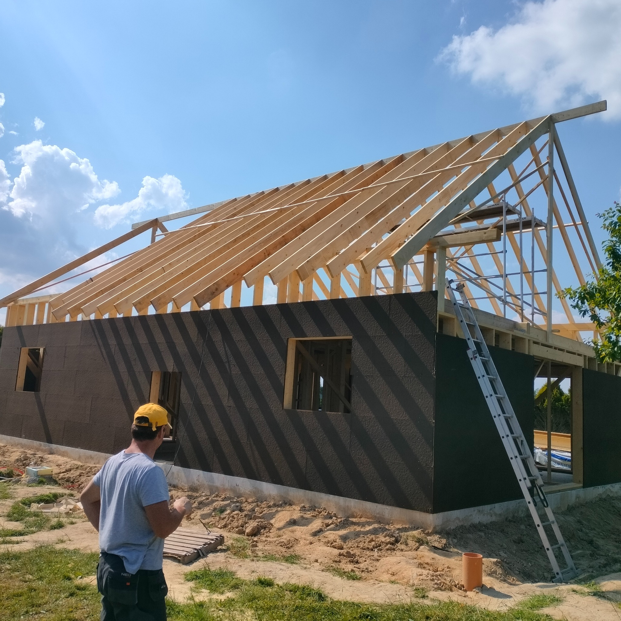 Budowa domu w technologii szkieletowej: widok na konstrukcję dachu z drewnianych krokwi, ściany pokryte ciemną płytą izolacyjną, pracownik w żółtej czapce i drabina oparta o ścianę.