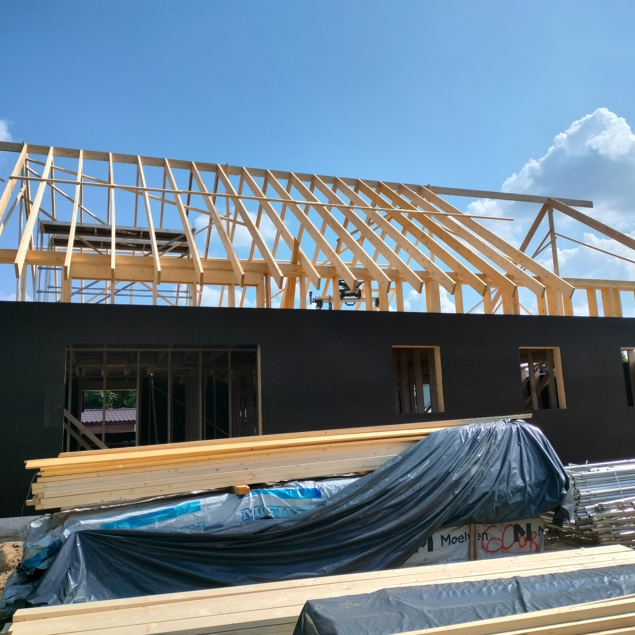 Konstrukcja szkieletowa domu w trakcie budowy, widoczne drewniane elementy więźby dachowej i fragment czarnej elewacji, z przodu stos drewna budowlanego przykryty folią z widocznym napisem 'Moelven'.