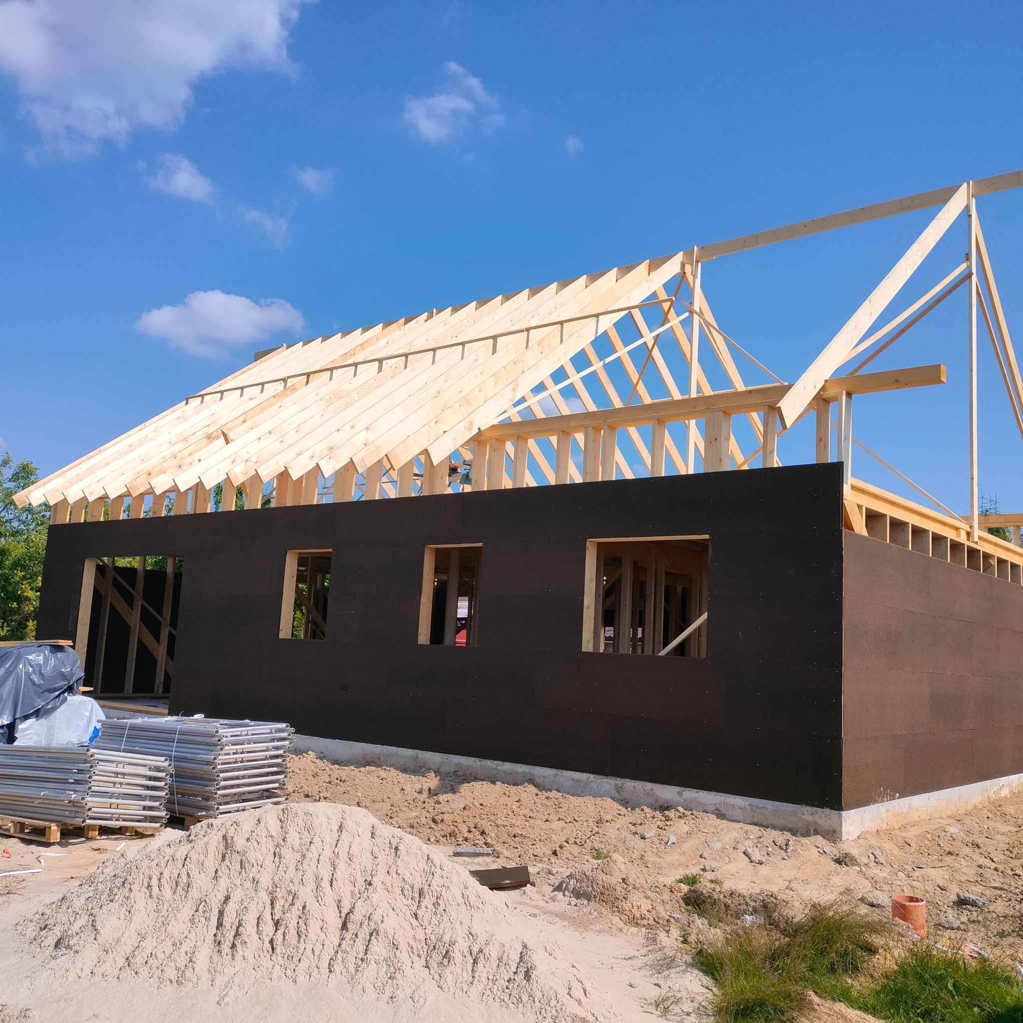 Surowy stan budynku z czarną elewacją i drewnianą konstrukcją dachu, widoczne rusztowania i piasek.