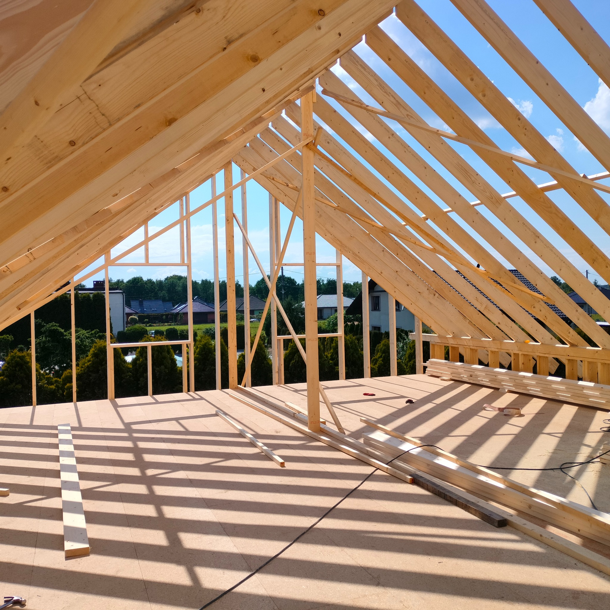 Konstrukcja drewniana dachu domu szkieletowego w trakcie budowy, widok na niebo i okoliczne domy przez otwory konstrukcji.