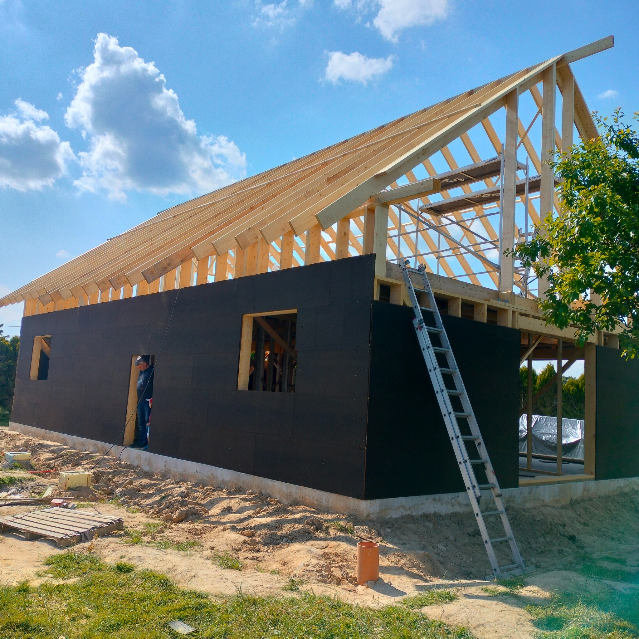 Budowa domu z drewnianą konstrukcją szkieletową, widoczne ściany z czarnej płyty, drabina oparta o ścianę, pracownik w drzwiach, błękitne niebo z chmurami w tle.