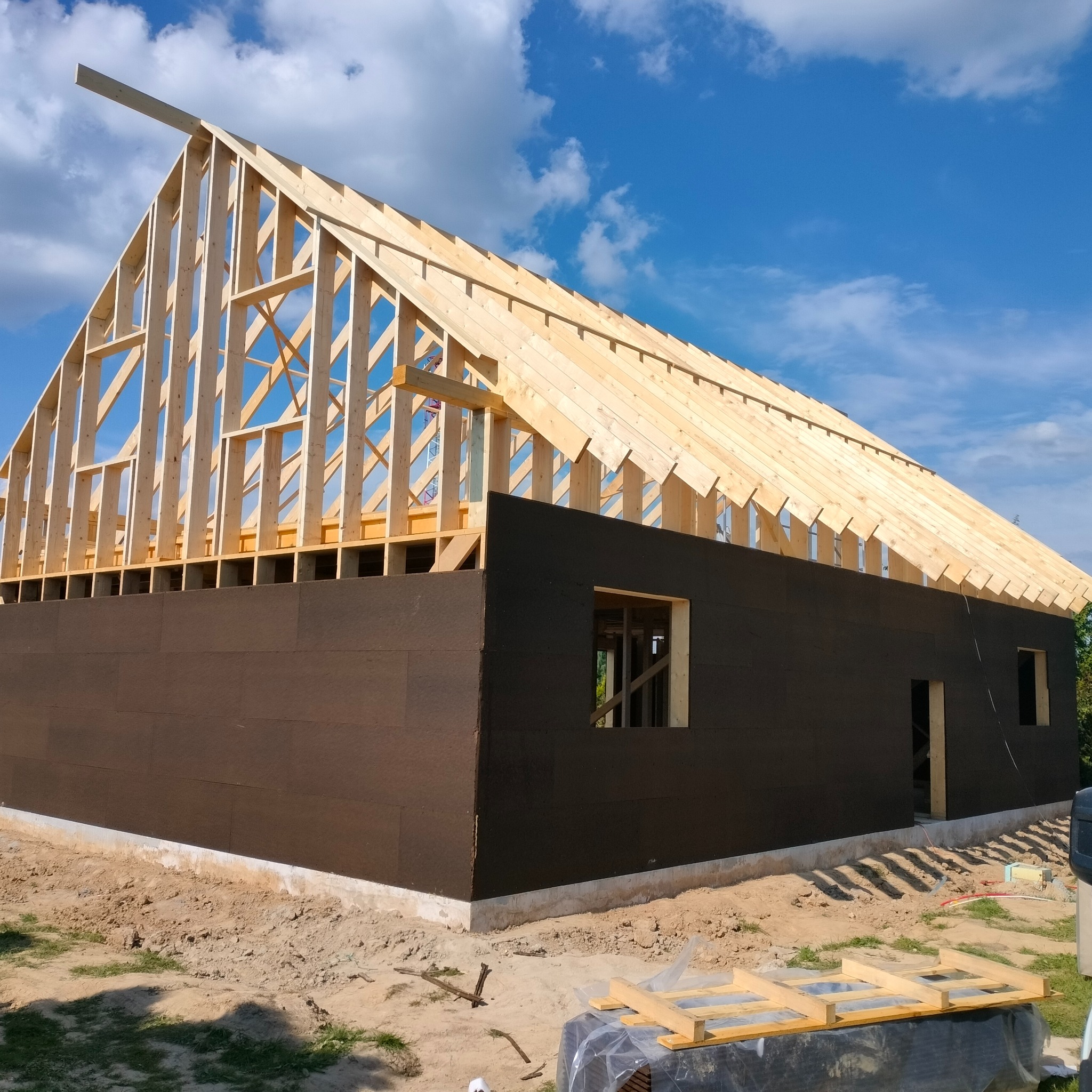 Konstrukcja domu w stanie surowym otwartym z drewnianym szkieletem i częściowo zamontowaną elewacją w Skrzeszewie Żukowskim, widoczne niebo z chmurami.