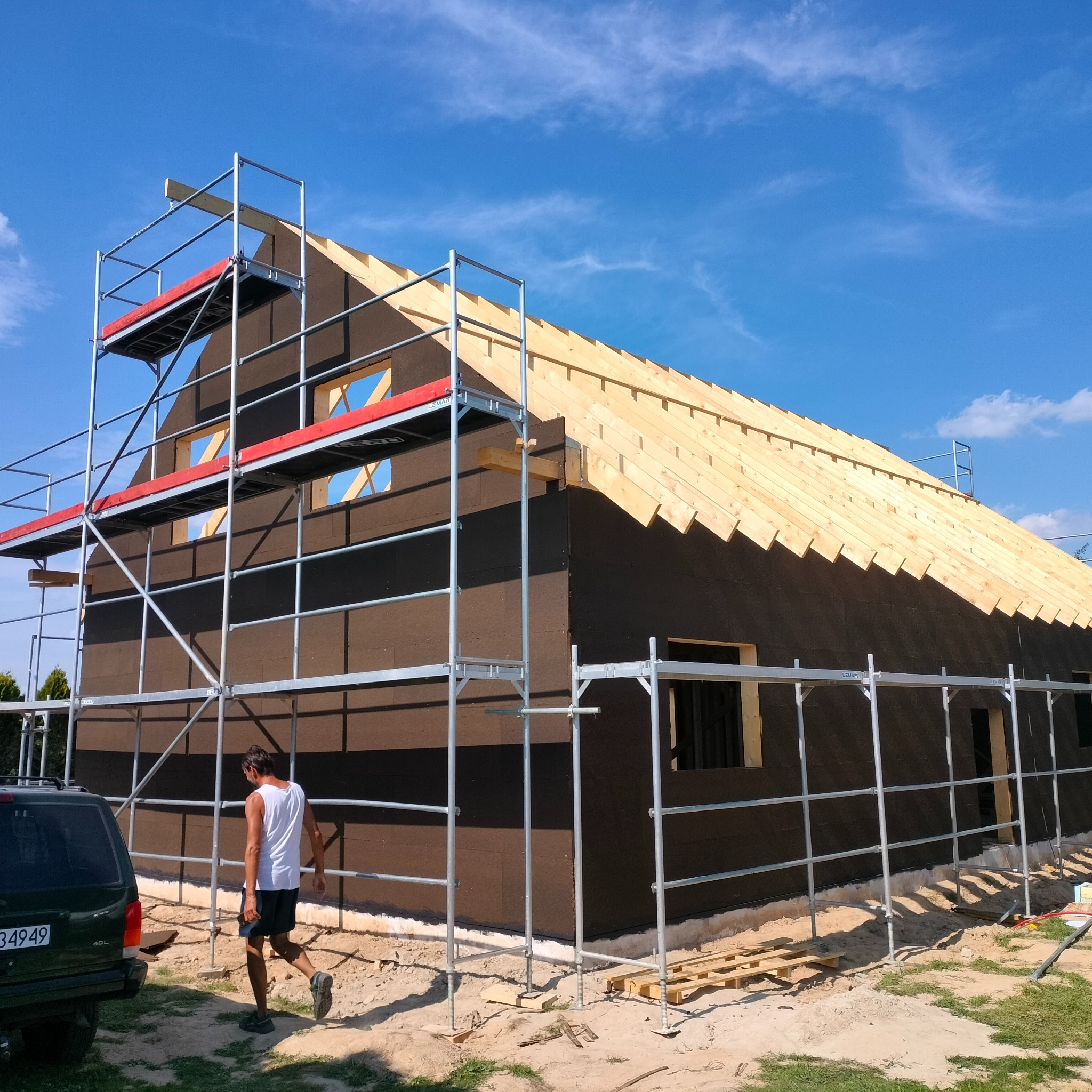 Budowa domu w technologii szkieletowej: widok na budynek z zewnątrz, z zamontowanymi rusztowaniami i częściowo wykonanym dachem z drewnianych krokwi, na tle błękitnego nieba. Mężczyzna w tle...