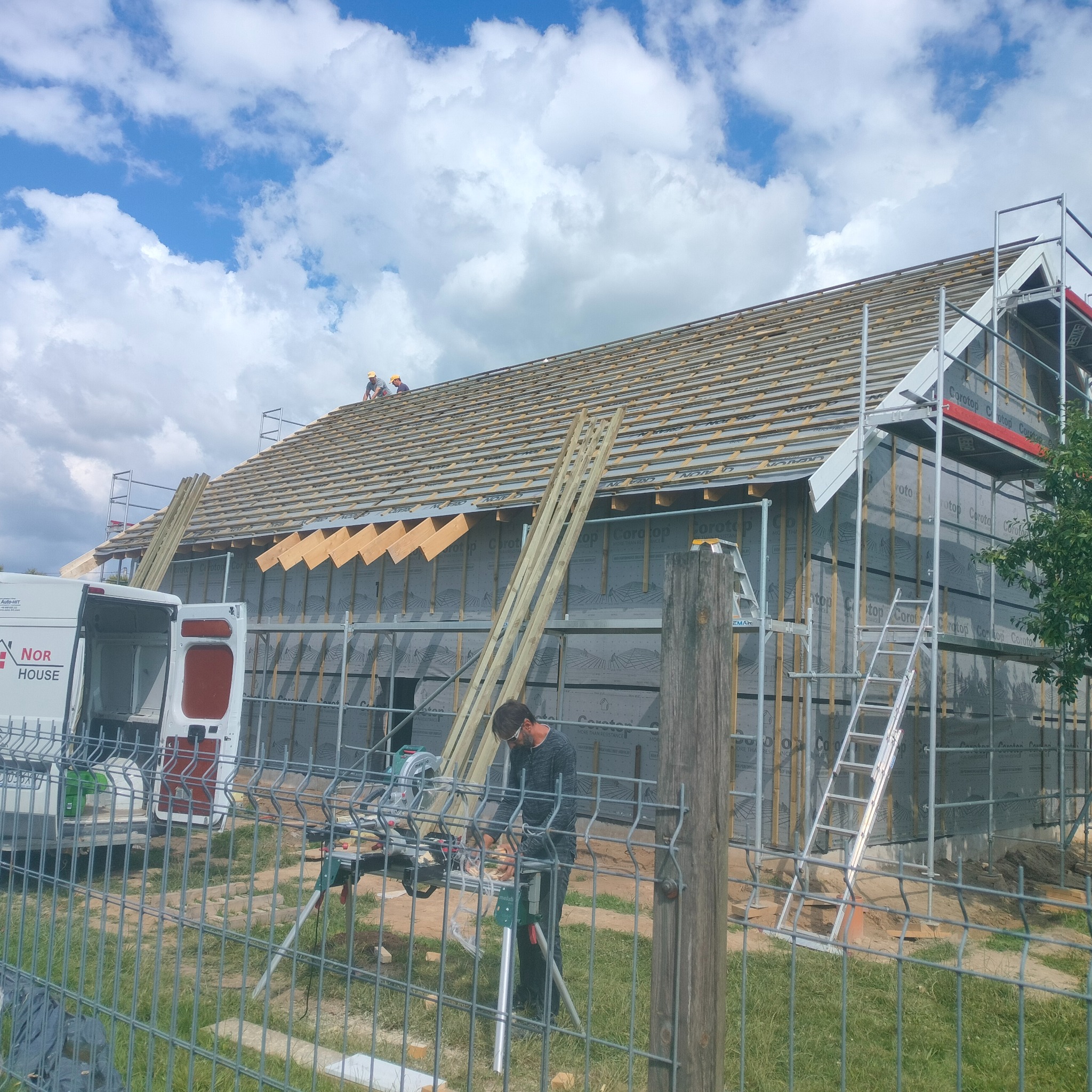 Budowa domu: pracownik tnie drewno na zewnątrz, w tle widoczny szkielet konstrukcji z izolacją, rusztowania i ekipa na dachu, częściowo zasłonięte przez metalowe ogrodzenie.