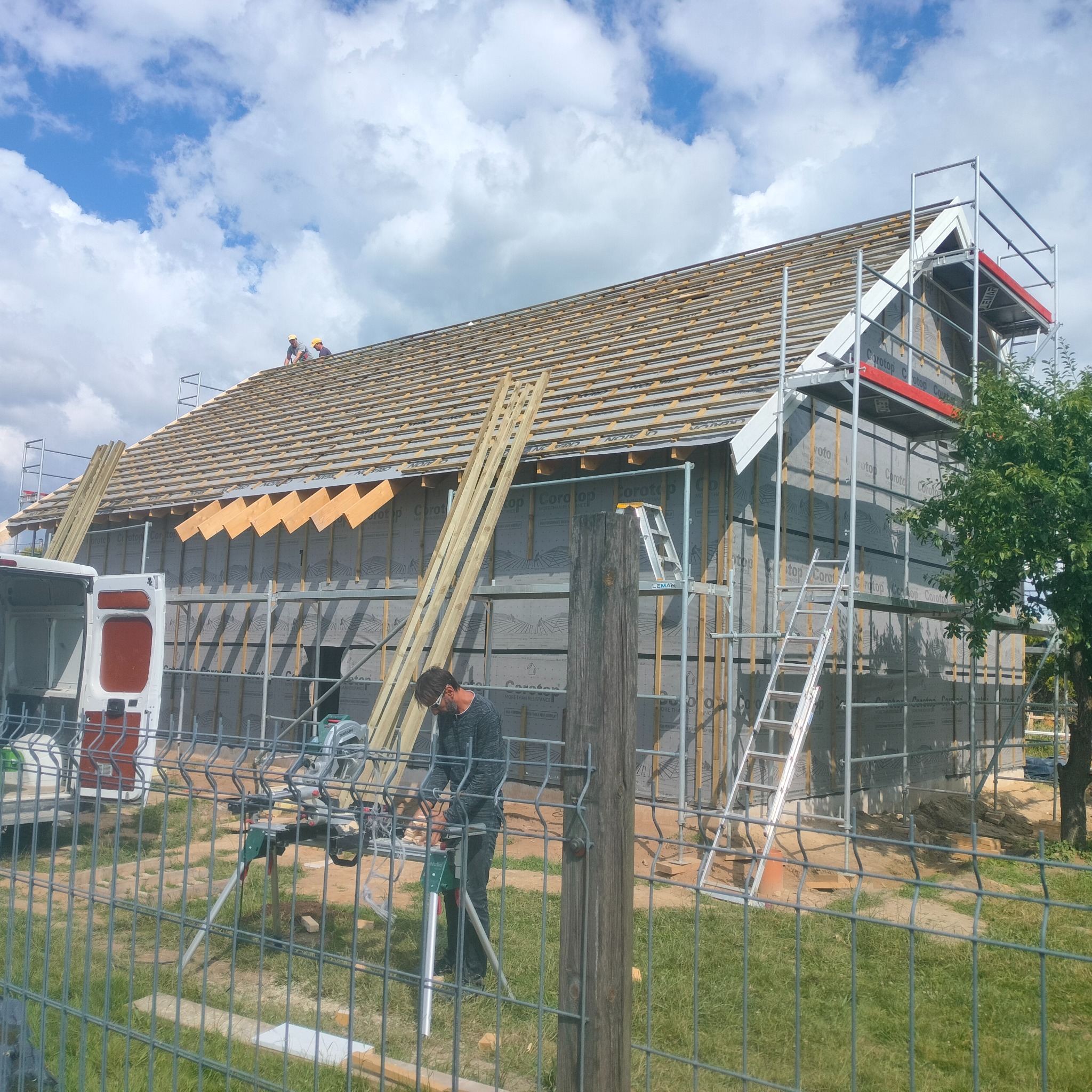 Budowa domu: robotnicy montują konstrukcję dachu i ocieplenie ścian zewnętrznych, widoczny ruszt, drabina i piła do cięcia drewna na pierwszym planie za ogrodzeniem.