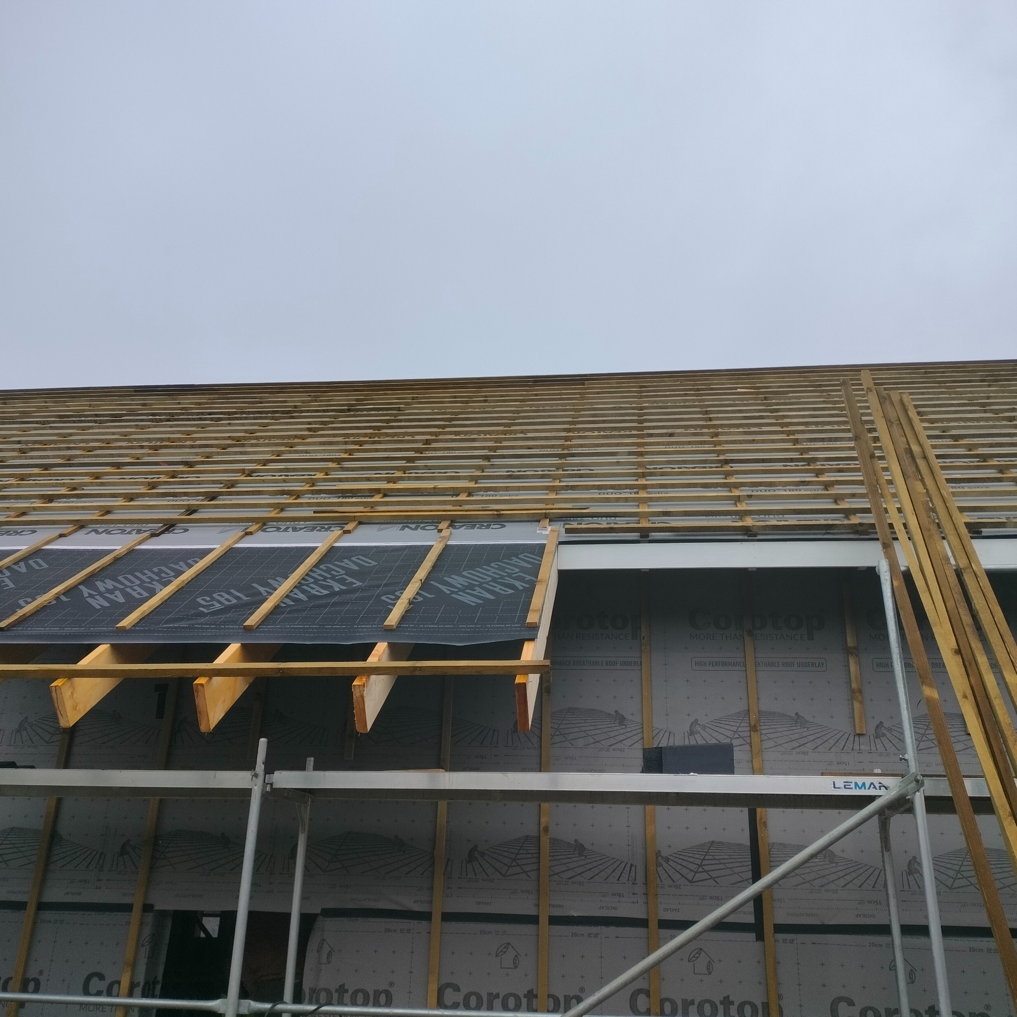 Konstrukcja dachu w trakcie budowy, widoczne drewniane elementy szkieletu, membrana dachowa z nadrukiem, rusztowanie LEMAN w dolnej części kadru, pochmurne niebo w tle.