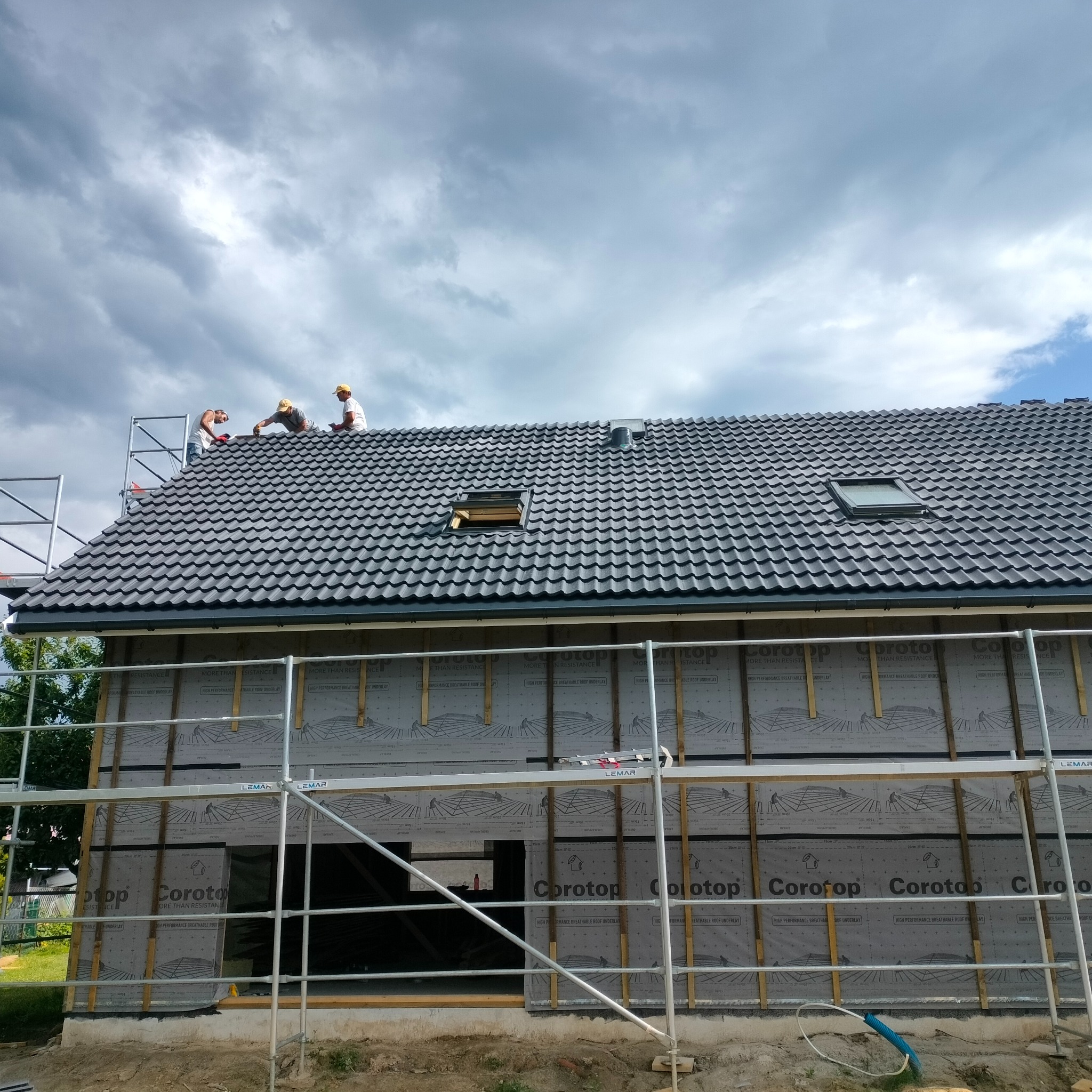 Budowa domu: ekipa dekarzy montuje dachówkę na dachu z oknami połaciowymi, widoczne rusztowania i membrana dachowa Corotop.