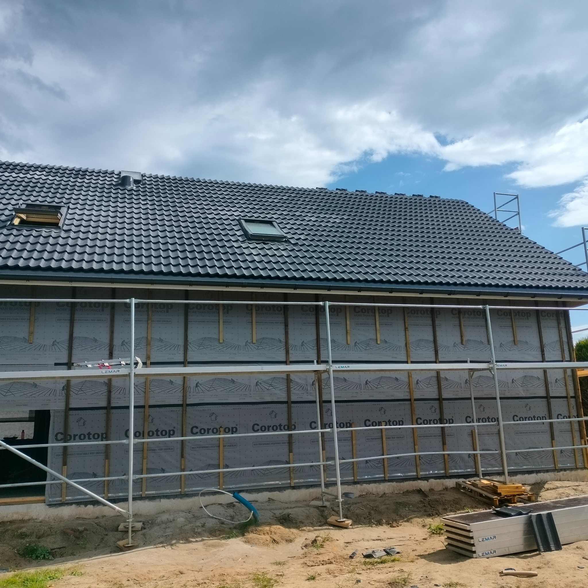 Budowa domu: widok na ścianę szkieletową z membraną Corotop i częściowo ułożonym dachem z dachówką ceramiczną, rusztowania dookoła.
