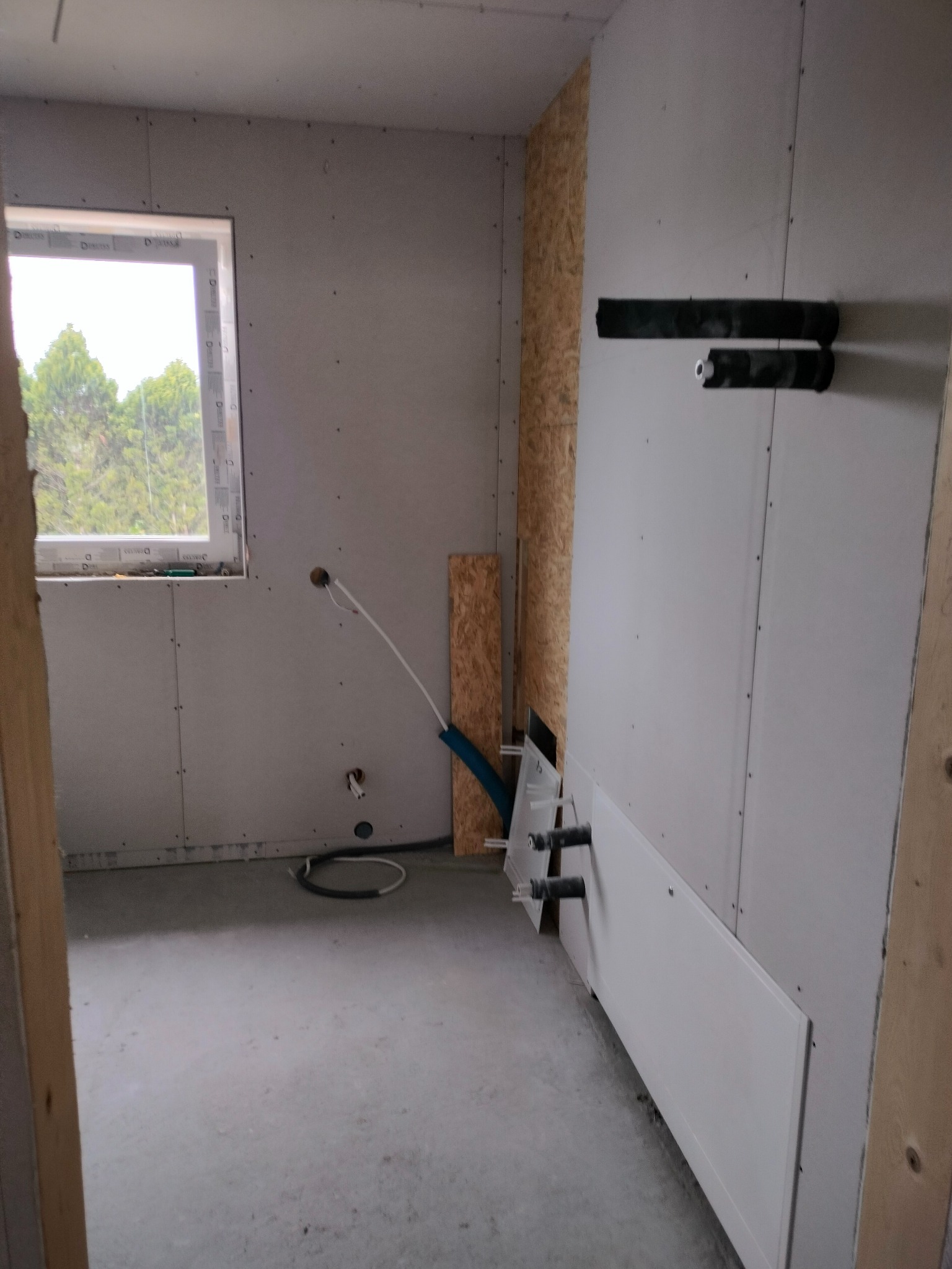 Wnętrze w trakcie budowy, widoczne ściany z płyt gipsowo-kartonowych, okno z widokiem na drzewa, instalacja wodno-kanalizacyjna w ścianie, surowa podłoga.