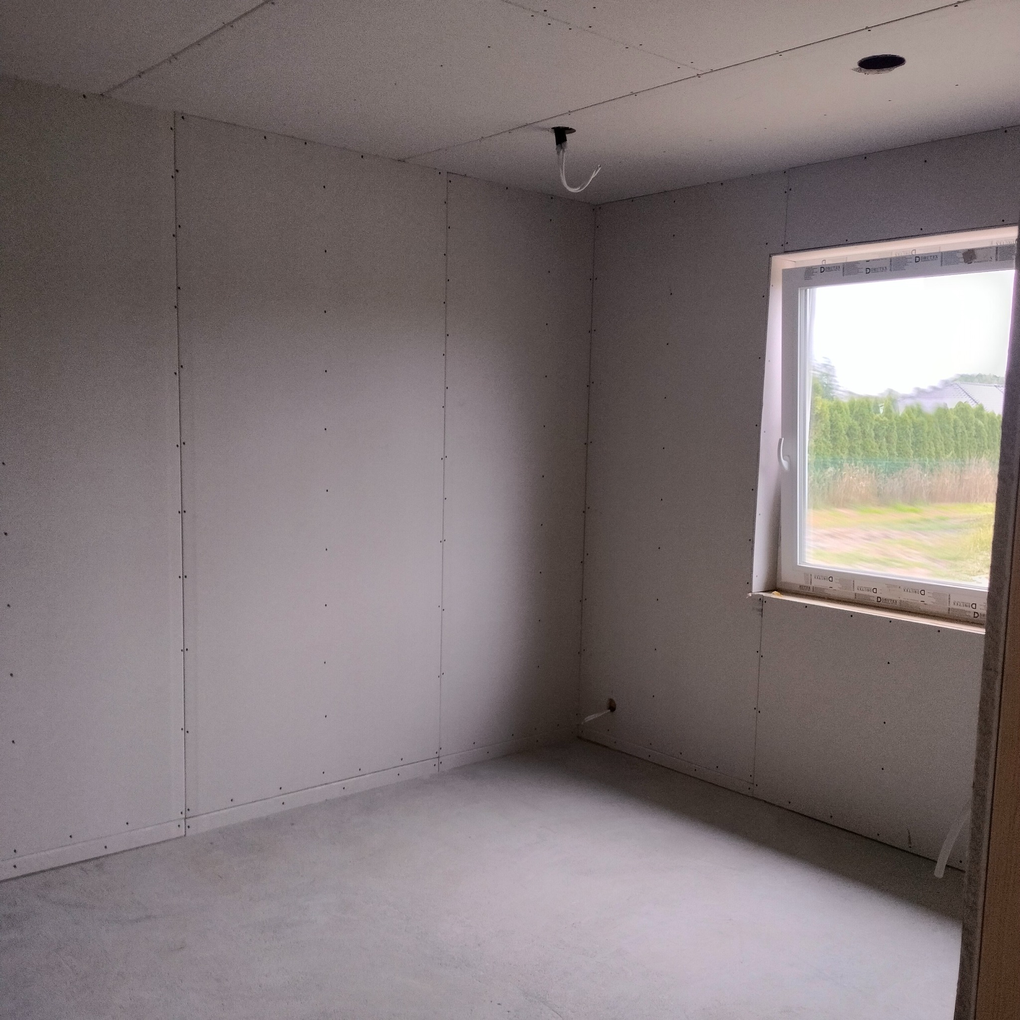 Wnętrze w stanie surowym: ściany wyłożone płytami gipsowo-kartonowymi, widoczne łączenia i wkręty, okno z widokiem na zieleń, betonowa posadzka, wyprowadzona instalacja elektryczna na suficie.