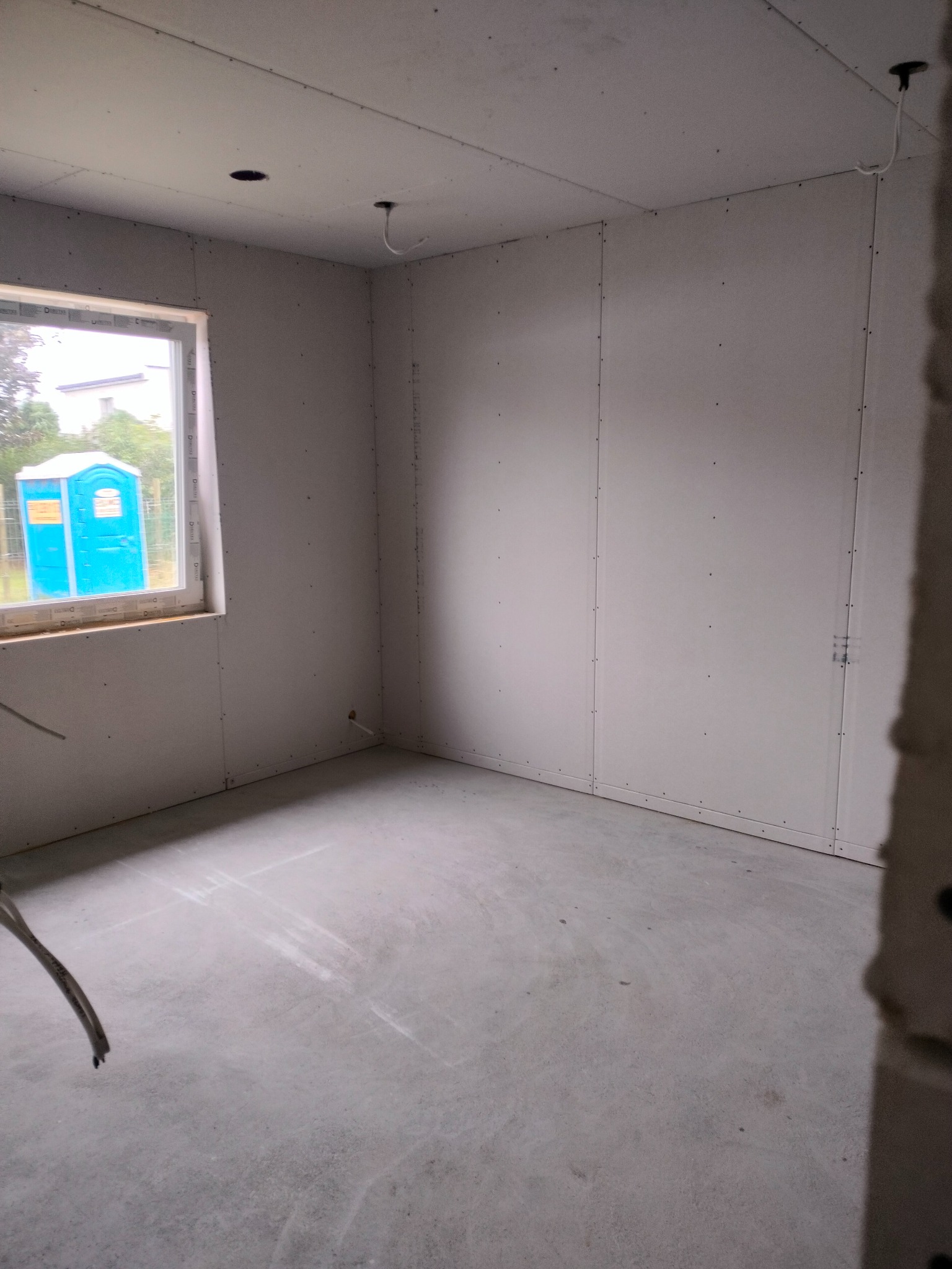 Wnętrze surowego pomieszczenia z oknem, ściany wykończone płytami gipsowo-kartonowymi, widoczna niebieska toaleta przenośna za oknem.