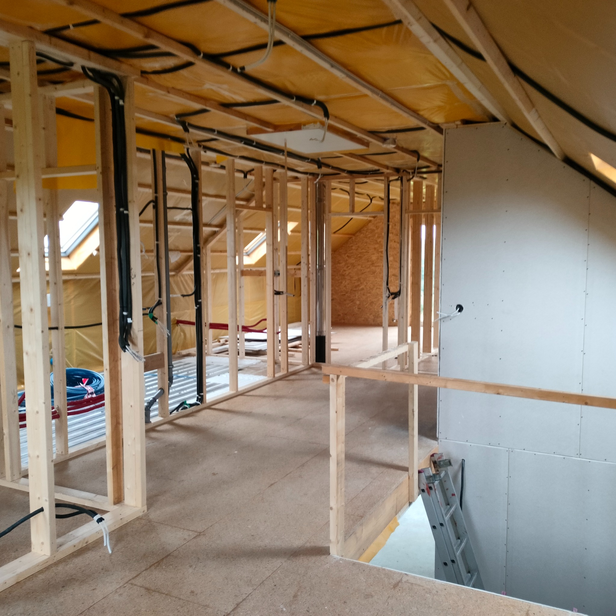 Wnętrze w trakcie budowy: drewniana konstrukcja szkieletowa, instalacje elektryczne i sanitarne, płyty gipsowo-kartonowe na poddaszu, widoczne okna dachowe i drabina.