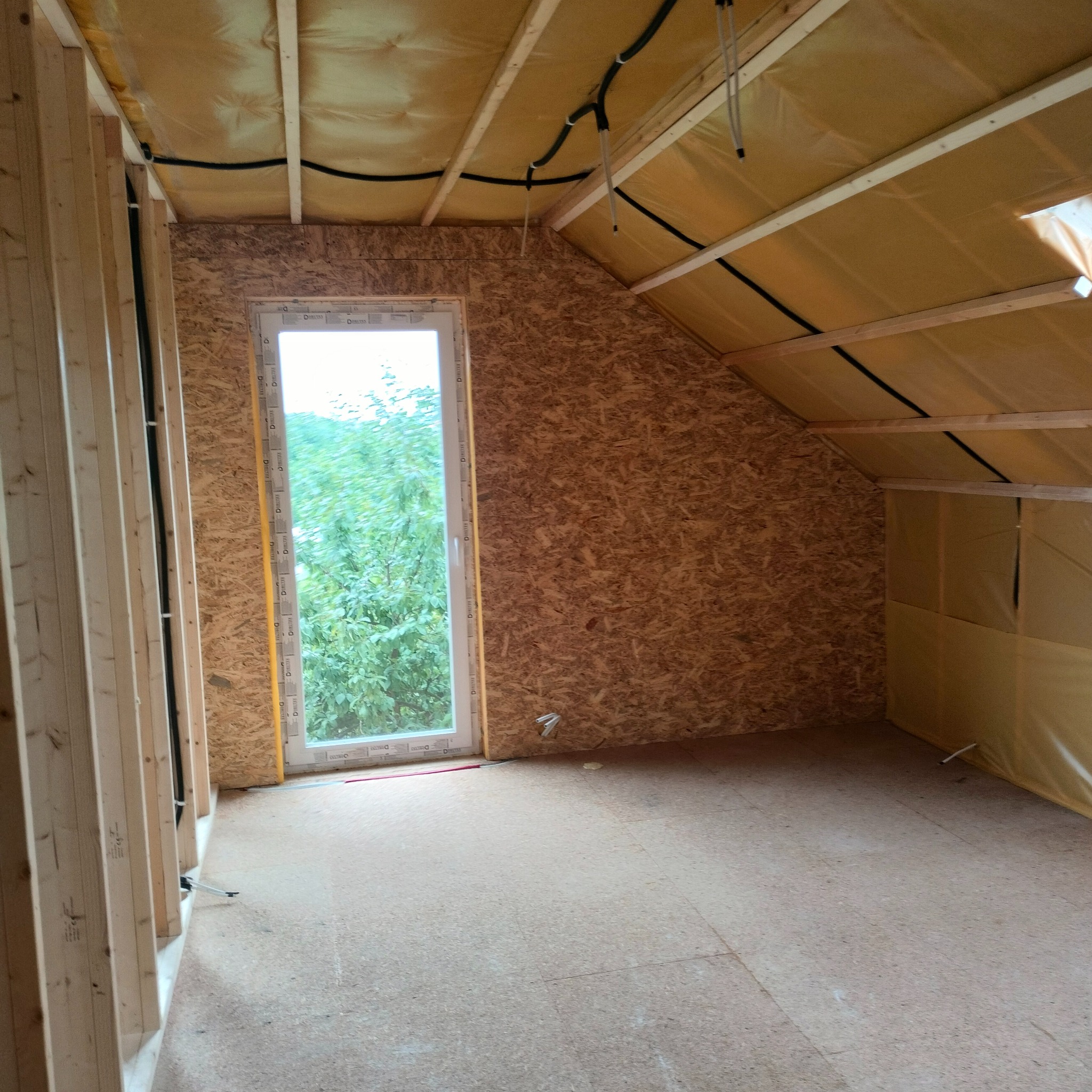 Wnętrze w trakcie budowy: szkielet drewniany, płyty OSB na ścianach i skośnym suficie poddasza, okno z widokiem na zieleń, instalacja elektryczna prowadzona po suficie.