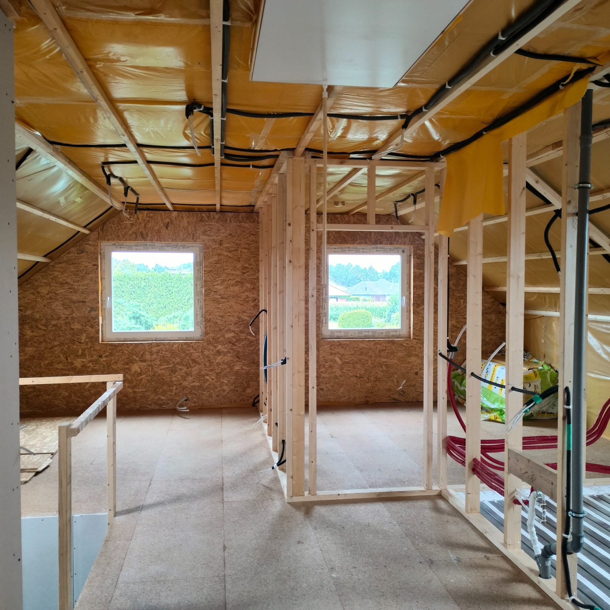 Wnętrze domu w trakcie budowy, widoczne drewniane ramy konstrukcyjne, izolacja stropu folią, okna z widokiem na zieleń, instalacje sanitarne i elektryczne w trakcie montażu.