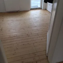 Jasna, odnowiona podłoga z desek w pokoju z grzejnikiem i oknem, widok z perspektywy progu drzwi.