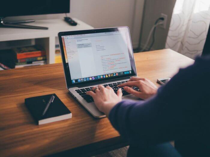 Osoba pisząca na laptopie na drewnianym biurku; obok leży zamknięty notatnik z długopisem i smartfon.