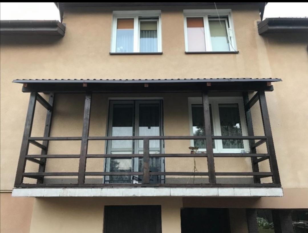 Zadaszony balkon z drewnianą konstrukcją i brązowym zadaszeniem, widok z zewnątrz na elewację budynku z dwoma oknami na górze.