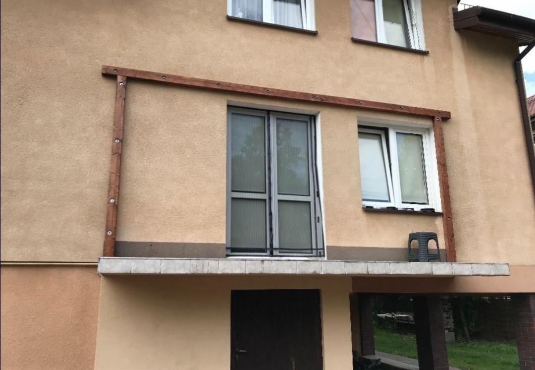 Montaż drewnianej konstrukcji, prawdopodobnie pergoli, przy ścianie budynku z oknami i balkonem, widoczne mocowania śrubowe.