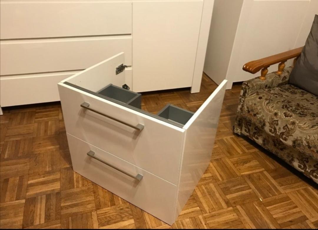 Biała, lakierowana szafka łazienkowa z dwiema szufladami i otwartą przestrzenią na umywalkę, widoczne szare wkłady szuflad, ustawiona na parkiecie w jodełkę, obok tapicerowany fotel.