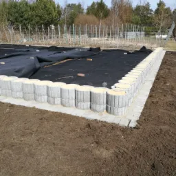 Przygotowanie podłoża pod taras: ułożona geowłóknina na ziemi, obrzeża z szarej kostki betonowej, widoczne elementy konstrukcji szklarni w tle.