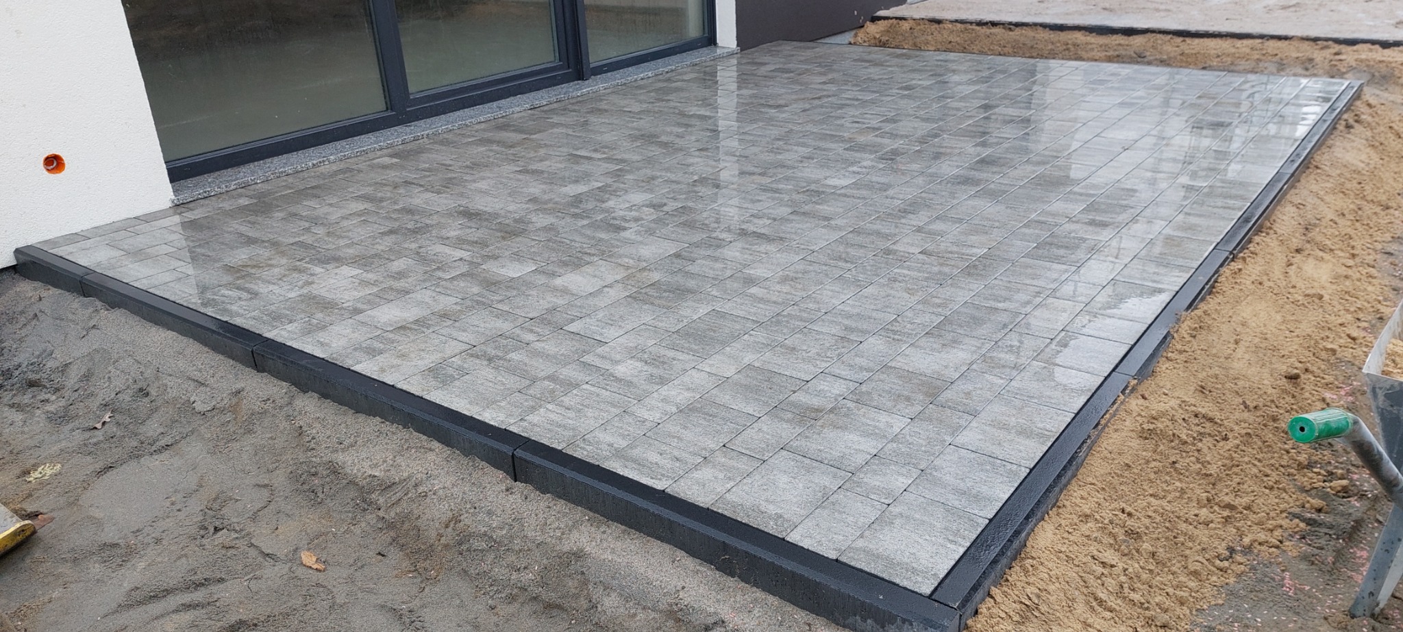 Świeżo ułożona, szara kostka brukowa na tarasie przylegającym do budynku, z czarnym obrzeżem, widoczna piaskowa podsypka i fragment taczki.