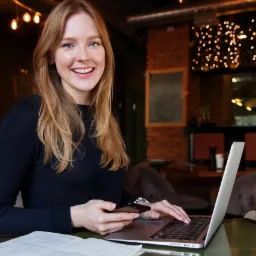 Uśmiechnięta blondynka w ciemnym swetrze pracuje na laptopie w kawiarni, trzymając telefon w dłoni i mając otwarty notatnik obok.